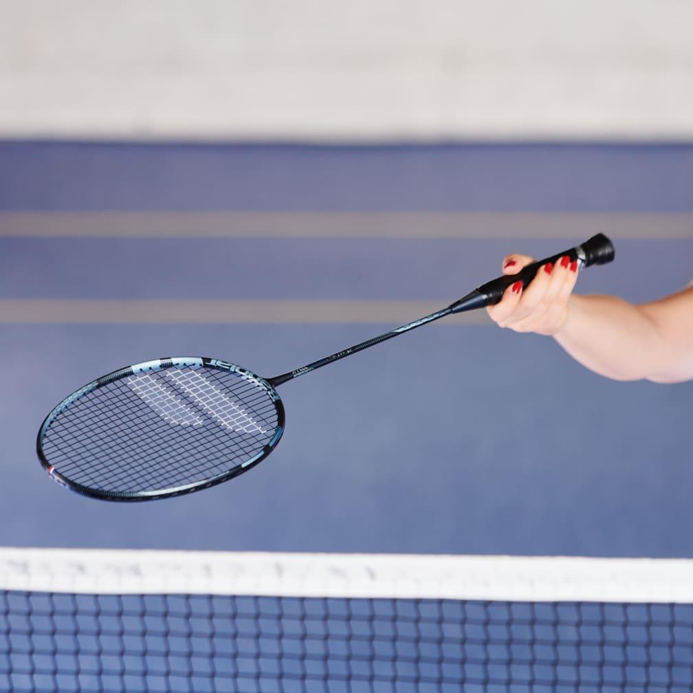 A person holding a Babolat X-Feel Essential Badminton Racket in Blue Grey, with a red-painted fingernail, displays its exceptional durability over the net on a blue court.