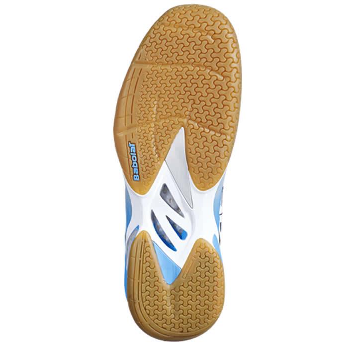 The image displays the sole of the Babolat Shadow Tour Badminton Shoes, characterized by a patterned tread for improved dynamic response. The light brown and white base includes a blue section with the "Babolat" label. This design appears to offer excellent grip, support, and shock absorption.