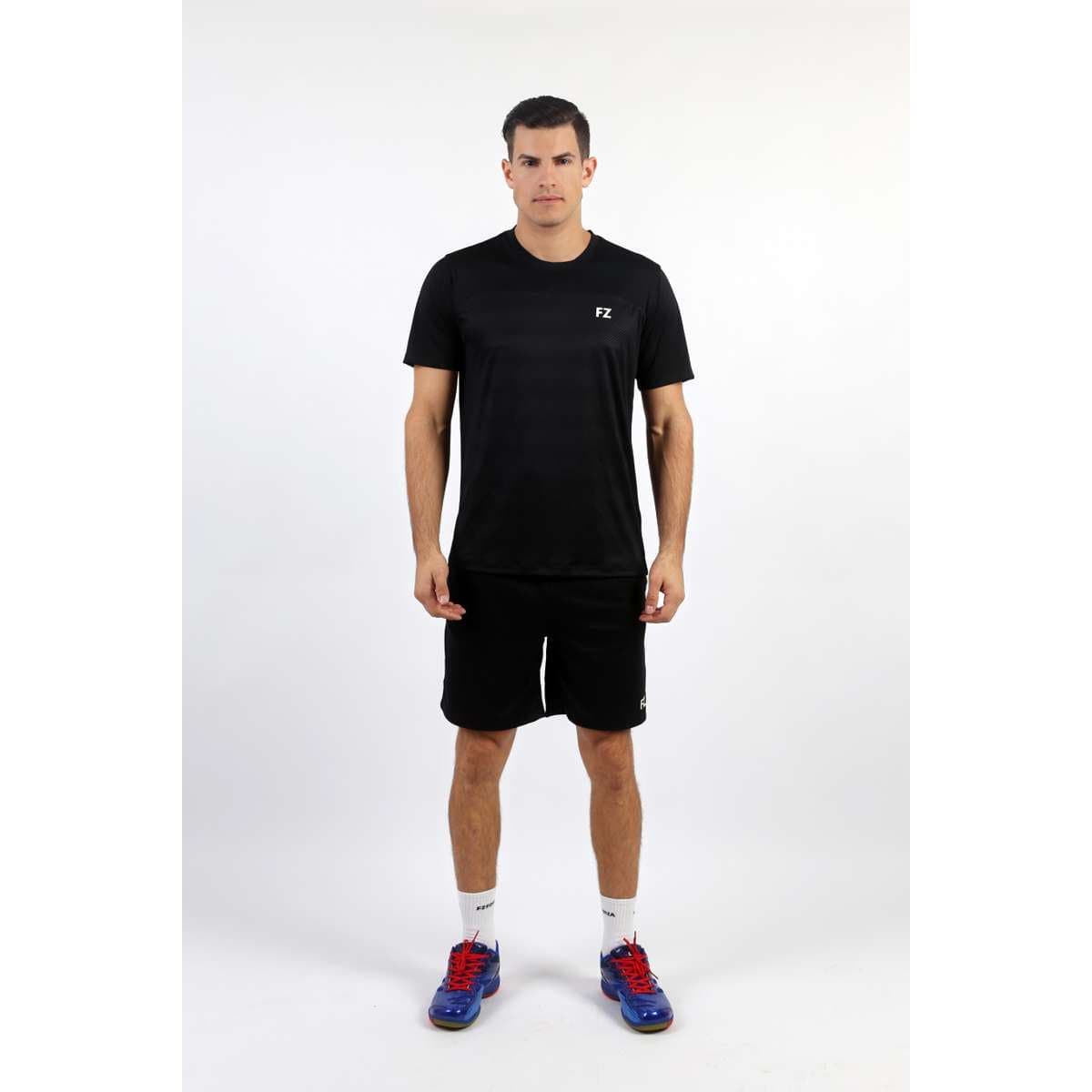 A man stands facing forward against a plain white background, wearing a black FZ Forza Helsinki Badminton T-Shirt with matching shorts, white socks, and blue athletic shoes with red laces.