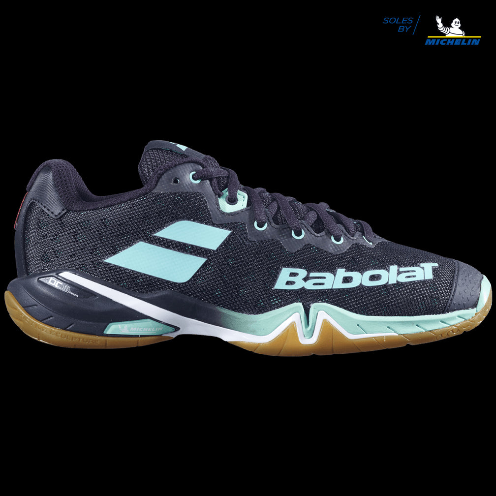 A pair of Babolat Shadow Tour badminton shoes in black with green accents, prominently displaying the Babolat logo and featuring a gum sole. The shoe showcases a textured design, visible laces, and integrates Michelin's sole technology for dynamic response with DCS technology.