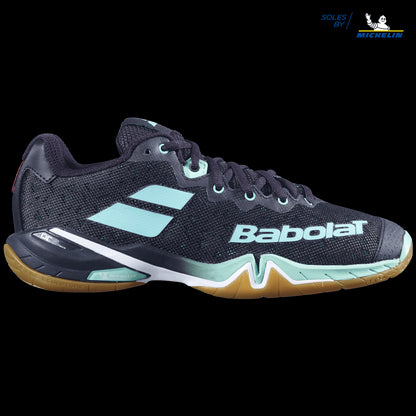 A pair of Babolat Shadow Tour badminton shoes in black with green accents, prominently displaying the Babolat logo and featuring a gum sole. The shoe showcases a textured design, visible laces, and integrates Michelin's sole technology for dynamic response with DCS technology.