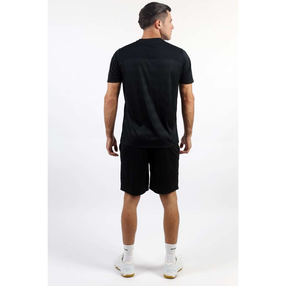 A man with short dark hair stands with his back to the camera, dressed in the FZ Forza Helsinki Badminton T-Shirt featuring DryForze technology and black shorts. He completes the look with white socks and sneakers against a plain white studio backdrop.