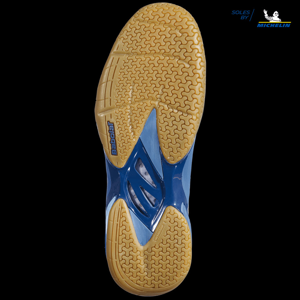 The image reveals the sole of a Babolat Shadow Spirit Badminton Shoe in dark blue, presenting a tread pattern with tan and blue sections. The "Soles by Michelin" logo is prominently displayed in the top right corner, emphasizing its incorporation of Michelin rubber as featured in Babolat's designs.