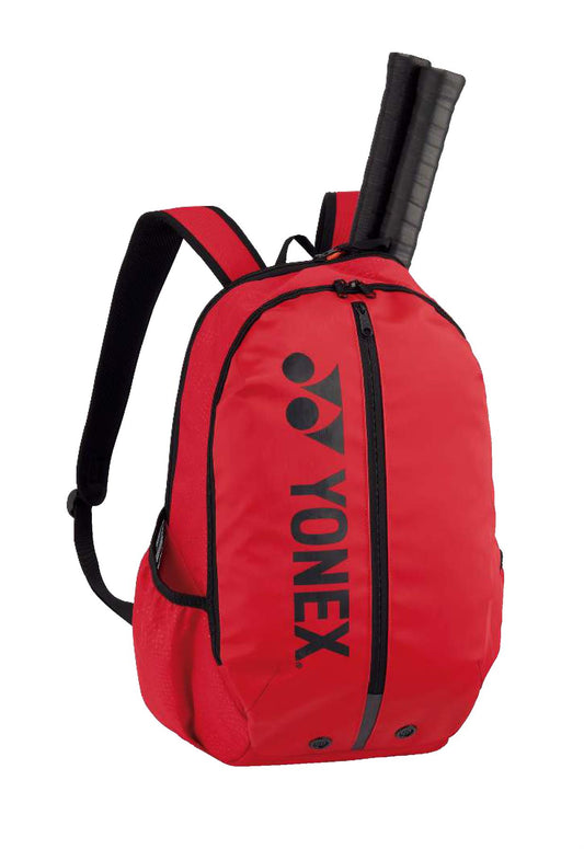 The Yonex Team Badminton Backpack 42012S in red, adorned with black zippers and straps, showcases the signature black logo on the front. Ideal for athletes, this durable backpack accommodates two tennis racket handles protruding from the top compartment.