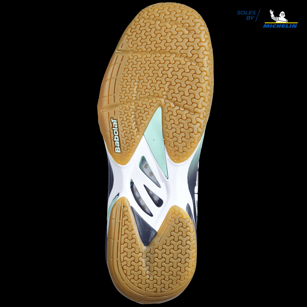 The image showcases the sole of a Babolat Shadow Tour Badminton Shoe, highlighting a dynamic response tread pattern designed for superior grip. The sole features a combination of light brown, white, and light blue on a black background, with the Michelin logo signifying DCS technology.