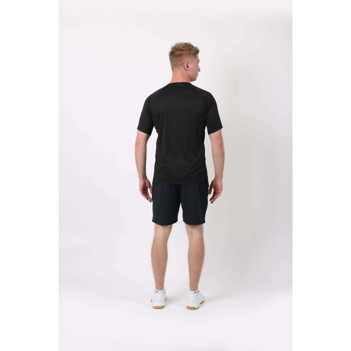 A person with short blonde hair is standing and facing away from the camera, showcasing the FZ Forza Forza Bling Men's Badminton T-Shirt in black. They wear dark shorts and white sneakers against a plain white background, ensuring breathability and style in every detail.