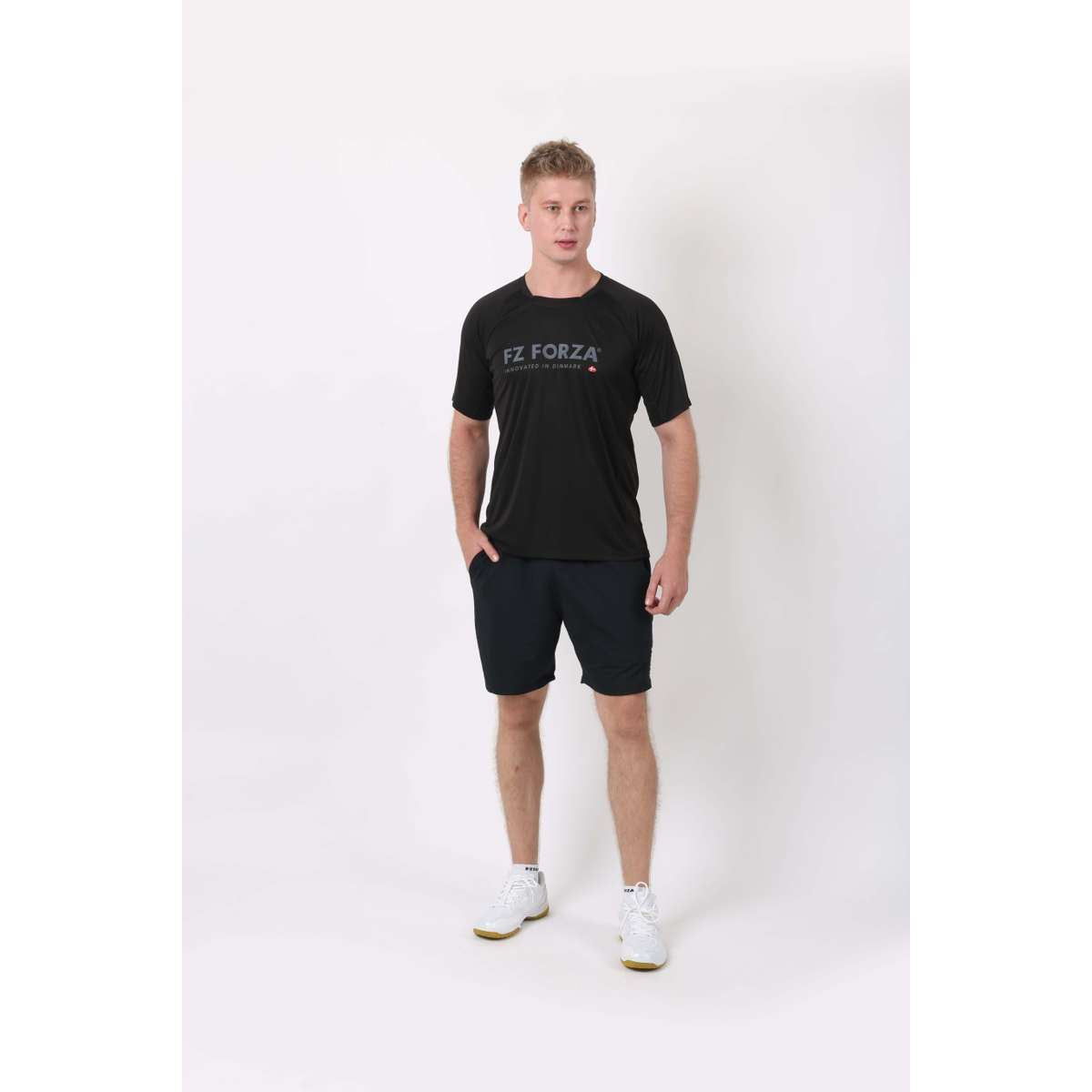 A man stands against a plain white background wearing an FZ Forza Forza Bling Men's Badminton T-Shirt in black, crafted from lightweight material. He pairs it with black shorts and white sneakers. With short hair and his hands in his pockets, he exudes a casual vibe while ensuring breathability.