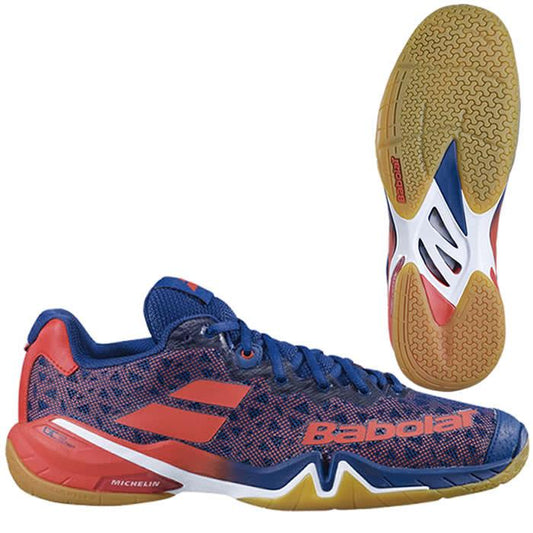 A pair of Babolat Shadow Tour Badminton Shoes in a black and orange design with gum-colored soles. The left shoe is shown in a side view, while the right shoe displays the sole pattern designed for optimal court grip.