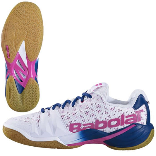 The Babolat Shadow Tour Badminton Shoes in White/Pink feature a white mesh upper with pink logo detailing and blue accents. The sole is a brown Michelin collaboration, featuring pink arch support and advanced DCS technology. The shoes are displayed from the side and bottom angles.