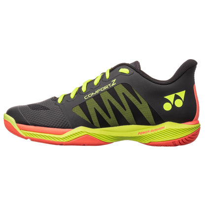 A black Yonex Power Cushion Comfort Z3 men's badminton shoe with a low profile design features neon yellow shades, lime green laces, and red accents. The brand's logo is prominently displayed on the side, while "Comfort Z," "Feather Bounce Foam," and "Power Cushion" emphasize its advanced sole and upper technology.