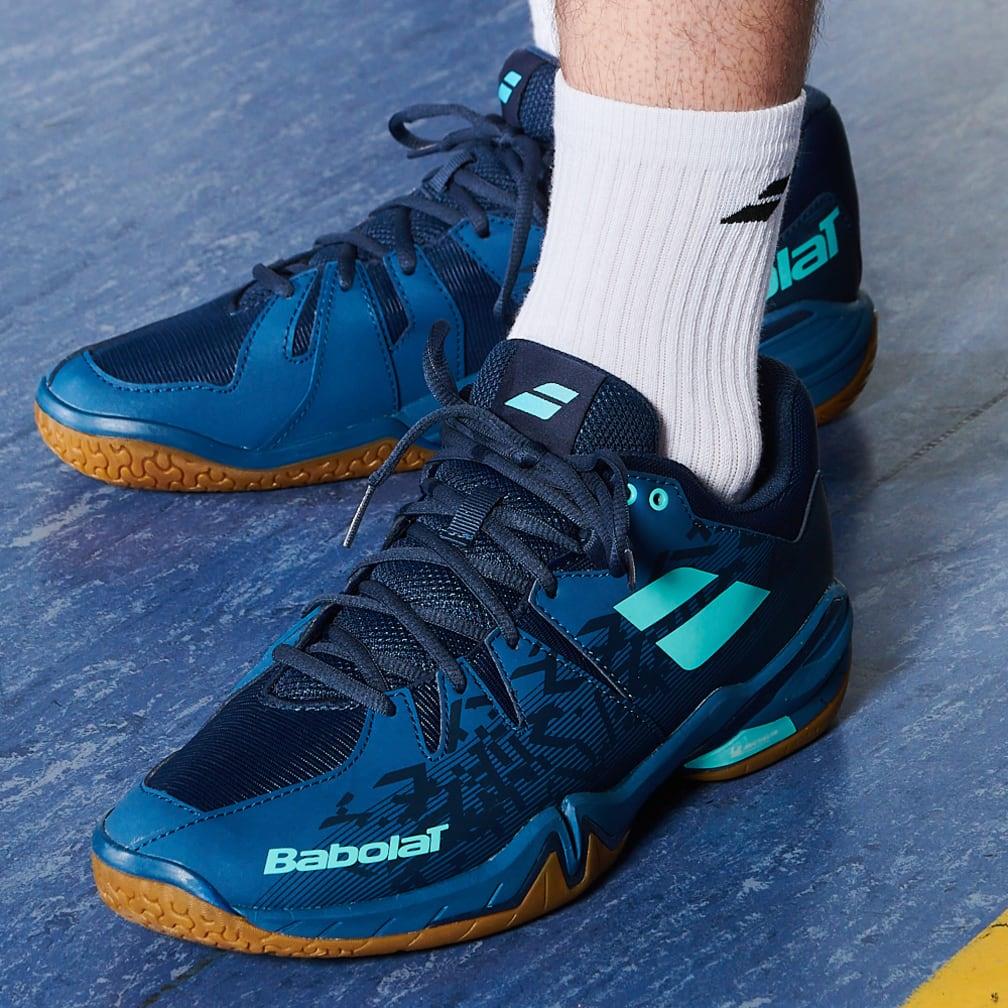 Close-up of a person wearing Babolat Shadow Spirit Badminton Shoes in Dark Blue with white socks on a blue surface. The shoes boast turquoise accents and a Michelin rubber gum sole, offering both style and durability.