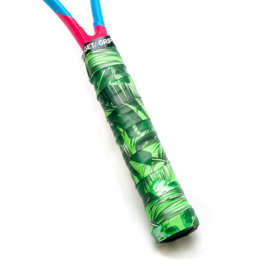 Close-up of a badminton racket handle wrapped with the Get A Grip Ace Tropic Green Overgrip, featuring a leaf-patterned design for excellent moisture absorption. The shaft above the non-slip grip is blue, and part of the pink racket frame is visible.