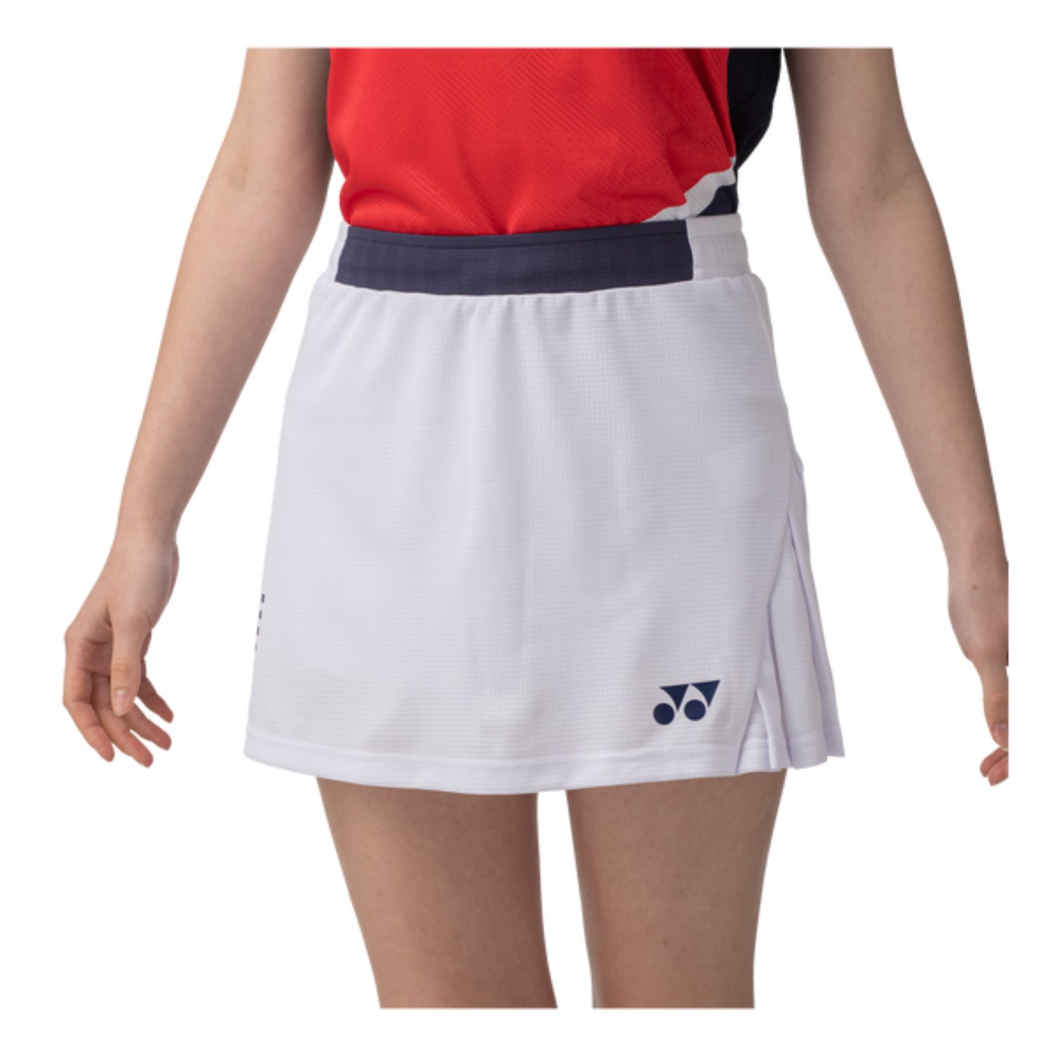 A person wearing a Yonex 26094 Womens Skort - White, paired with a red and black top. The skort includes a navy waistband, UV reduction technology, and is adorned with a small logo on the bottom front. Their hands rest by their sides against a neutral white background.