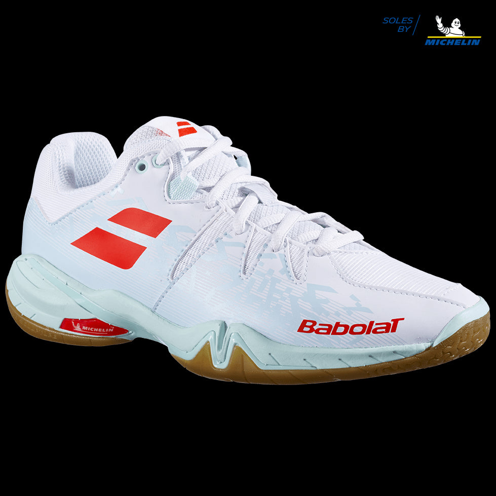 The Babolat Shadow Spirit Women's Badminton Shoes in white and light blue showcase the iconic red Babolat branding on the side and top. These shoes feature a gripsole designed with "Soles by Michelin" on a black background for exceptional traction, complemented by a lace-up design to ensure a secure fit.